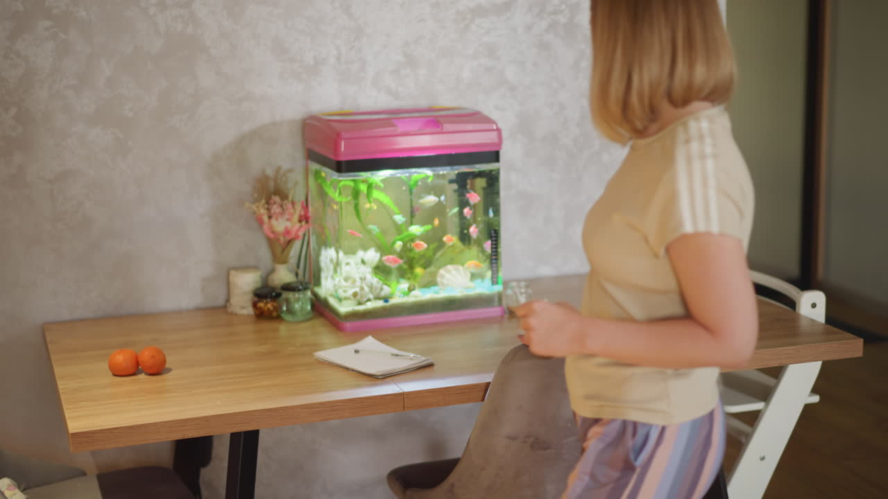 Woman in casual outfit with striped pants walks indoors near pink aquarium filled with colorful fish and green plants, creating cozy home interior with modern decor and peaceful relaxed atmosphere