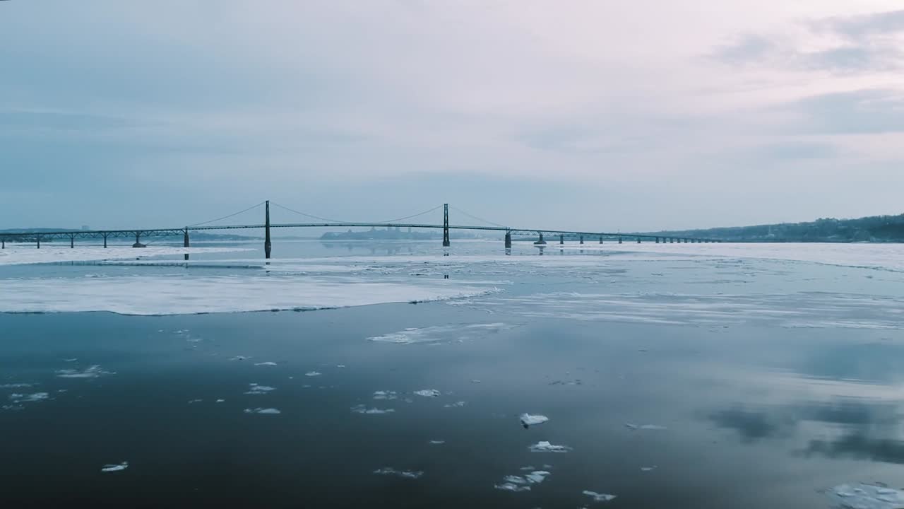 Suspension bridge stretches across ice fringed waters on Saint Lawrence River near Quebec City as winter landscape forms calm scene captured from aerial drone view with soft blue tones