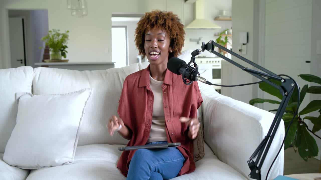 African American woman addressing camera into boom mic on sofa at home prompted by tablet