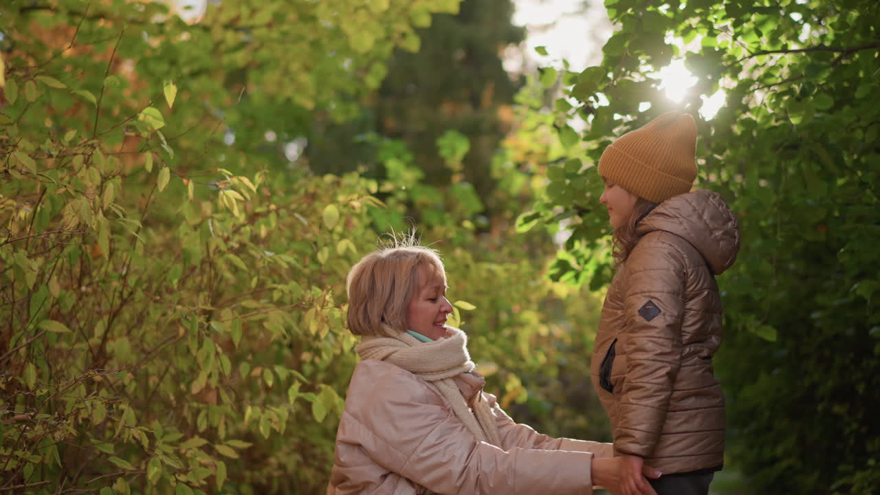 lovely moment with mother straightening daughter hoodie in sunlit autumn woods, warm light filtering through green and yellow foliage, expressions tender and joyful