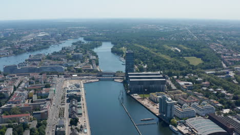 Aerial-tilt-down-and-pan-view-Treptower-building-complex-and-Molecule-Man-sculpture-on-Spree-river.-Berlin,-Germany