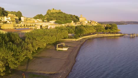 beautiful drone shot along lake bracciano coast reveals historic town center, fortress at sunset