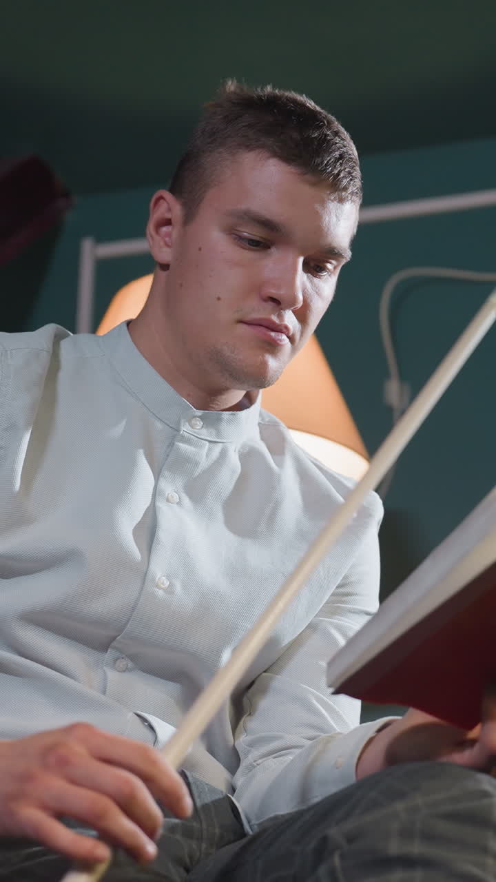 joven con camisa blanca y pantalones grises sosteniendo un libro rojo y un palo de señalización. la expresión pensativa sugiere un enfoque profundo. la iluminación cálida mejora la atmósfera intelectual y estratégica
