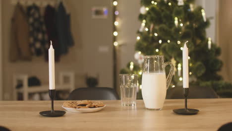 camera focuses on an empty glass, a jug of milk and a plate full of cookies on an empty table with two candles in a room decorated with a christmas tree