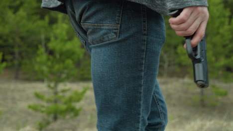 a close view of a man wearing a gray jacket and blue jeans, holding a handgun by his side, walking through a forest path