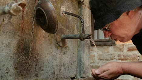 Man-wearing-hat-and-glasses-cupping-hands-and-drinking-cold-water-to-quench-thirst-from-an-old-village-well-spring-water-supply-fountain