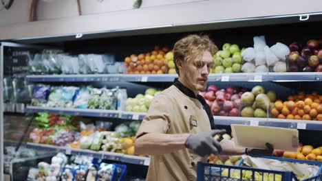 hombre trabajador con delantal negro y guantes almacenando las frutas en el supermercado. joven empleado en el trabajo. lado