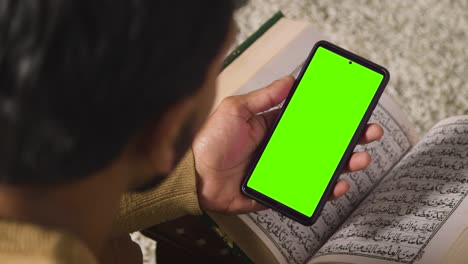 Close-Up-Of-Muslim-Man-At-Home-Sitting-On-Floor-And-Reciting-From-The-Quran-Holding-Green-Screen-Mobile-Phone