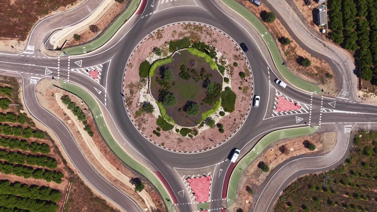 Top-down view of a large roundabout with a green landscaped central island, multiple entry roads, road markings, and sparse vehicle traffic in a rural area
