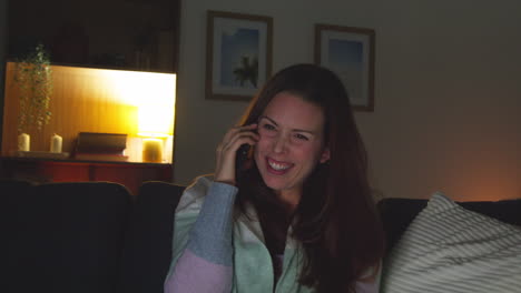 smiling woman sitting on sofa at home at night talking on mobile phone and watching movie or show on laptop 5