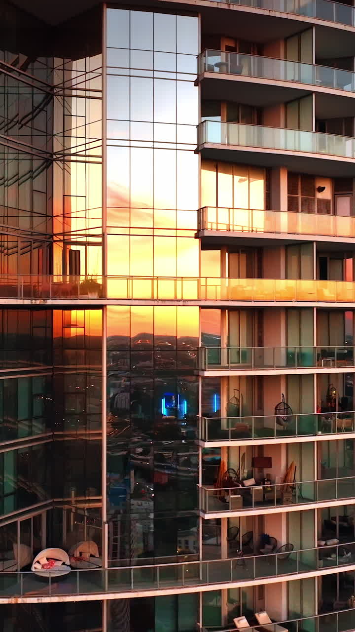 Mirror windows of modern multi-storied high-rise block of flats reflecting beautiful light at sunset. Drone flying around the building at sundown. Vertical video