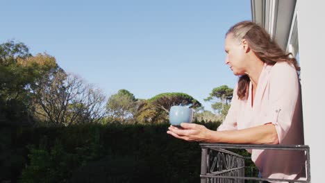 Thoughtful-senior-caucasian-woman-standing-on-sunny-balcony-drinking-coffee-and-looking-around