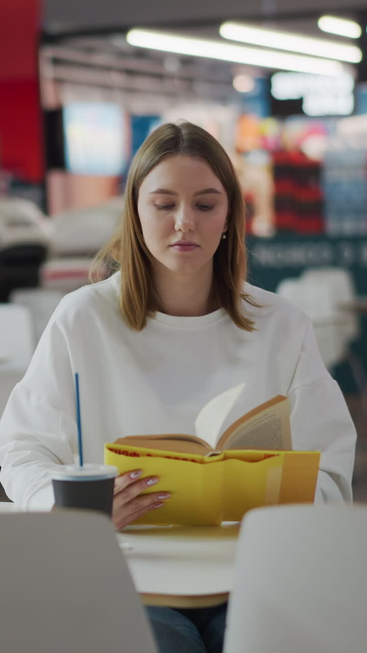 White lady seated in a mall reading a book with a coffee cup and shopping bag nearby, soft lighting creates a relaxed atmosphere with blurred background