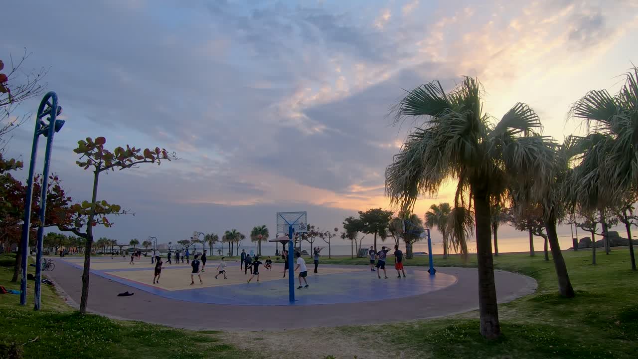Teenagers play pick up games of basketball at ocean side courts with evening sunset in background.