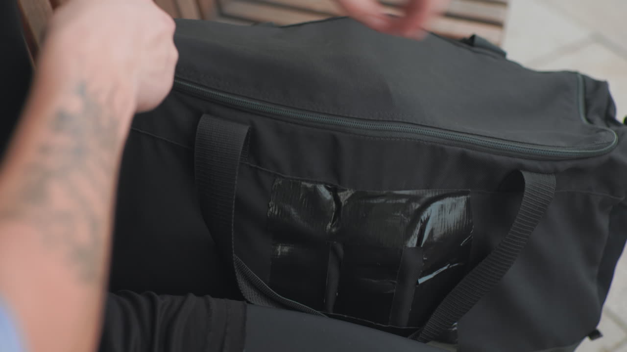 Uomo bianco che chiude la cerniera di una borsa da viaggio nera, allaccia la cinghia e fissa un guantone da boxe all'interno, avambraccio tatuato visibile, routine post-allenamento ordinata sulla panca, atmosfera da pendolare urbano, pronto per andare al prossimo impegno