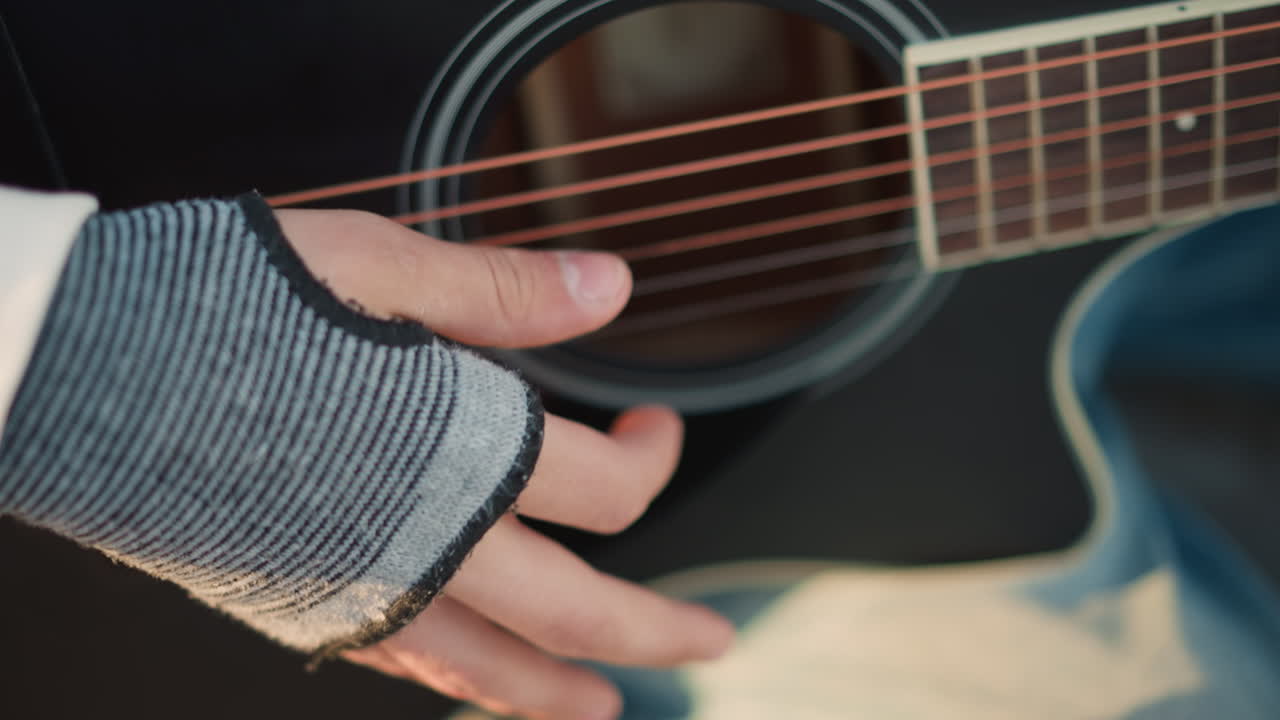 primo piano di un chitarrista bianco che pizzica una chitarra nera, messa a fuoco sulla buca con guanto grigio senza dita, ambiente interno intimo, risonanza tonale calda, corde e rosetta dettagliate, atmosfera rilassata da prova