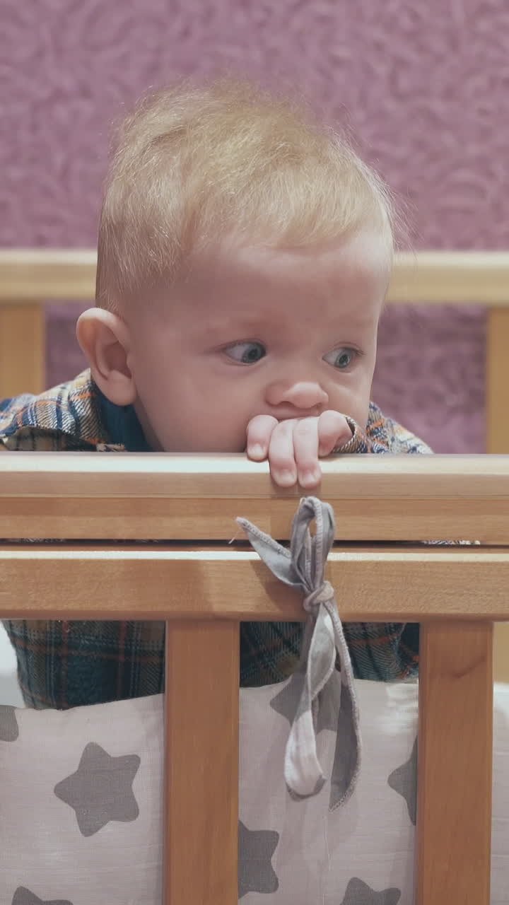 funny little boy leans on edge of wooden cot with star patterned linen against pink wall in light room closeup