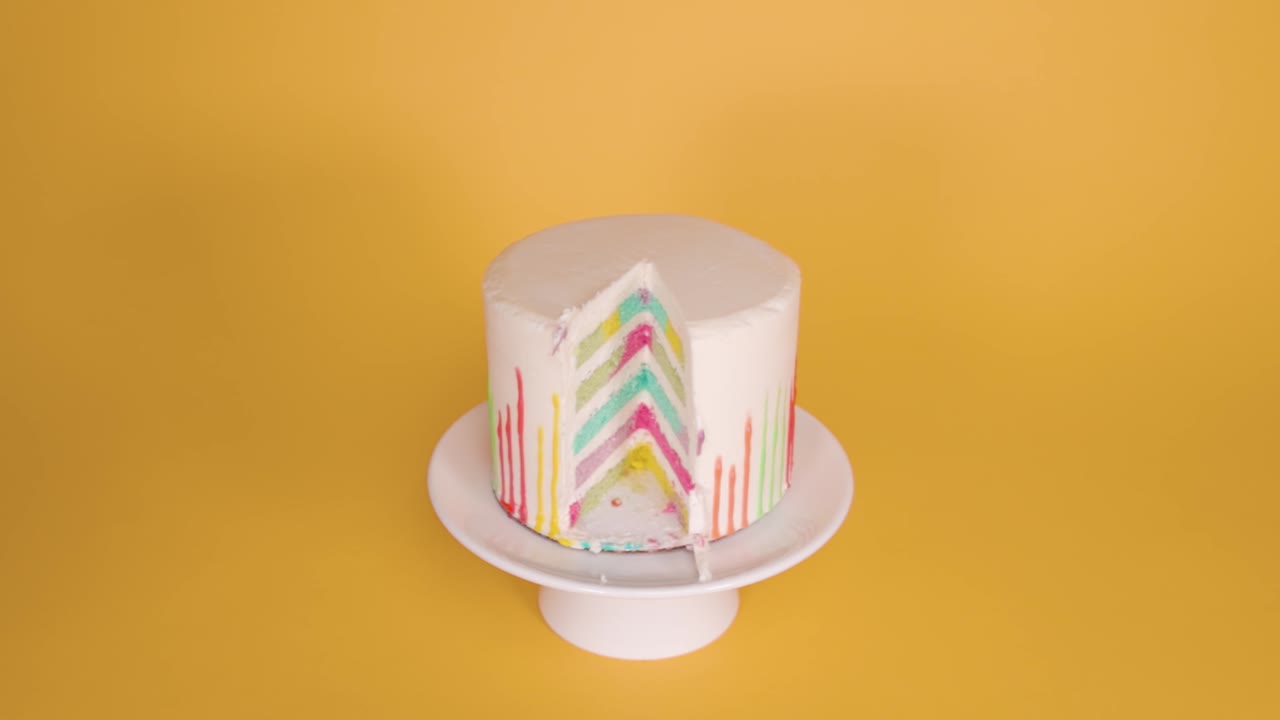 A vibrant slice of rainbow cake being lifted from a plate, set against a solid yellow backdrop