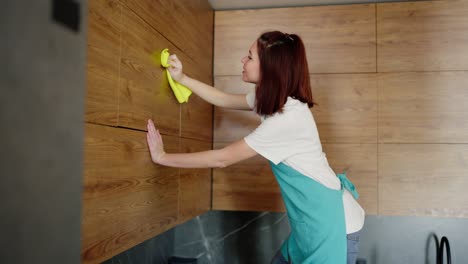 Side-view-of-a-confident-brunette-cleaning-lady-girl-in-a-white-t-shirt-and-blue-apron-wiping-the-upper-cabinets-in-a-modern-kitchen-with-a-wooden-texture-using-a-yellow-rag-while-cleaning-a-studio-apartment-on-call