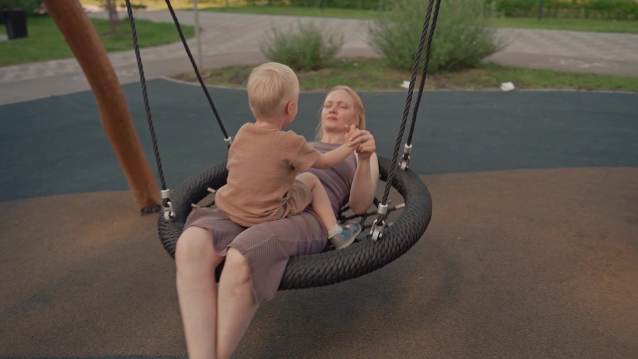 Donna e bambino condividono un momento tenero su un'altalena a pneumatico al parco. Il bambino siede tra le sue gambe tenendosi per mano mentre dondolano dolcemente insieme in un ambiente di parco giochi all'aperto con erba verde.