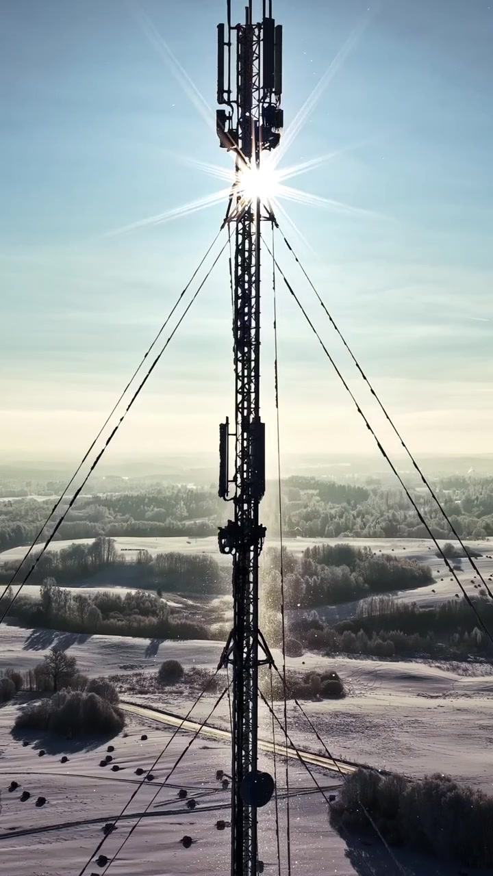 Torre di telecomunicazioni che si erge sopra la campagna innevata sotto un sole splendente, silhouette verticale di drone retroilluminata.