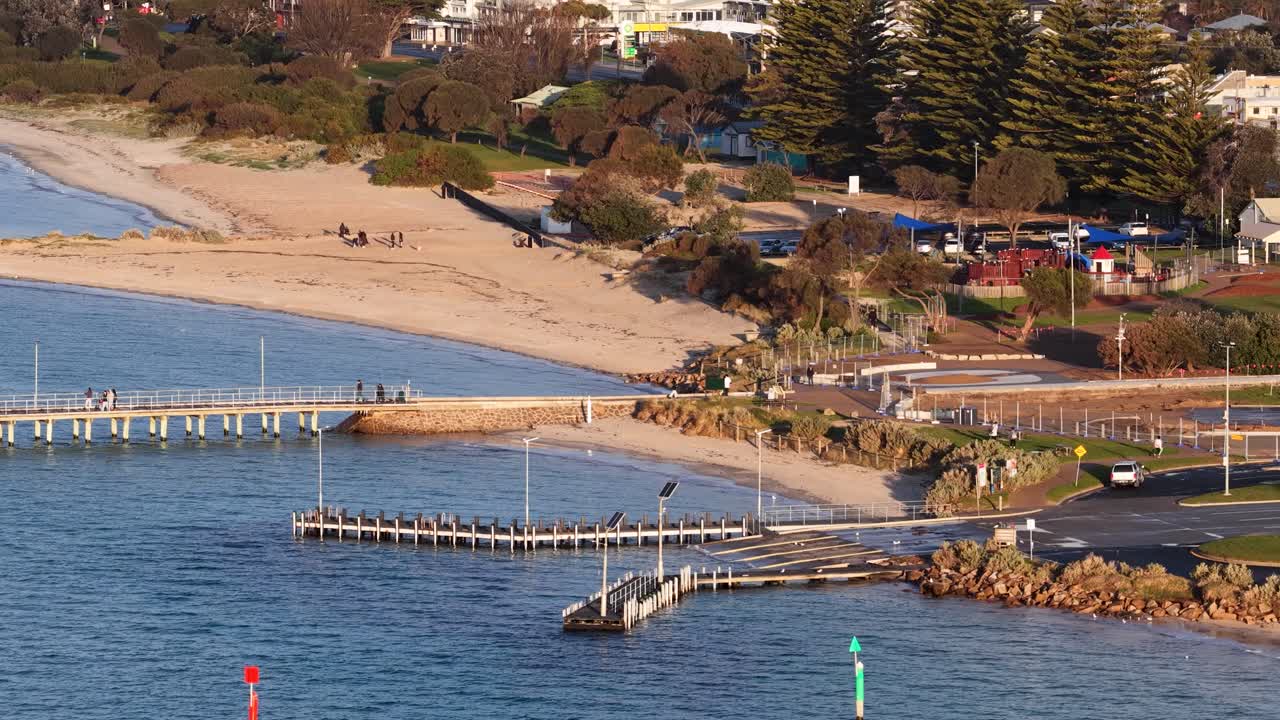 Aerial camera pans across Portsea Pier, sandy beach, and coastal landscape in warm evening light