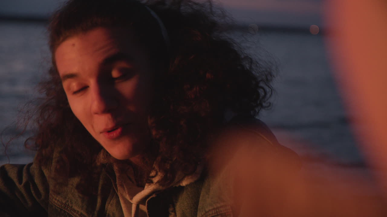 Camera Focuses On A Teenage Boy Smiling Sitting Around Bonfire With Her Friends