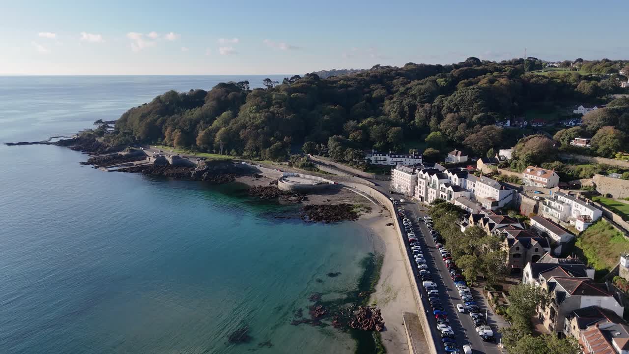 213 Séquence de drone à haute altitude au-dessus de la baie de Havelet, Saint Peter Port, Guernesey, montrant une baie calme avec une mer claire, des bassins de baignade et des vues sur les falaises boisées sous le soleil de fin d'après-midi avec un ciel sans nuages.