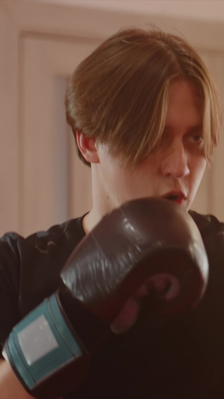 Giovane uomo con capelli castani corti che indossa guantoni da boxe neri. I primi piani si concentrano sui movimenti della parte superiore del corpo. L'illuminazione brillante evidenzia la forma del soggetto durante i movimenti di pugno.