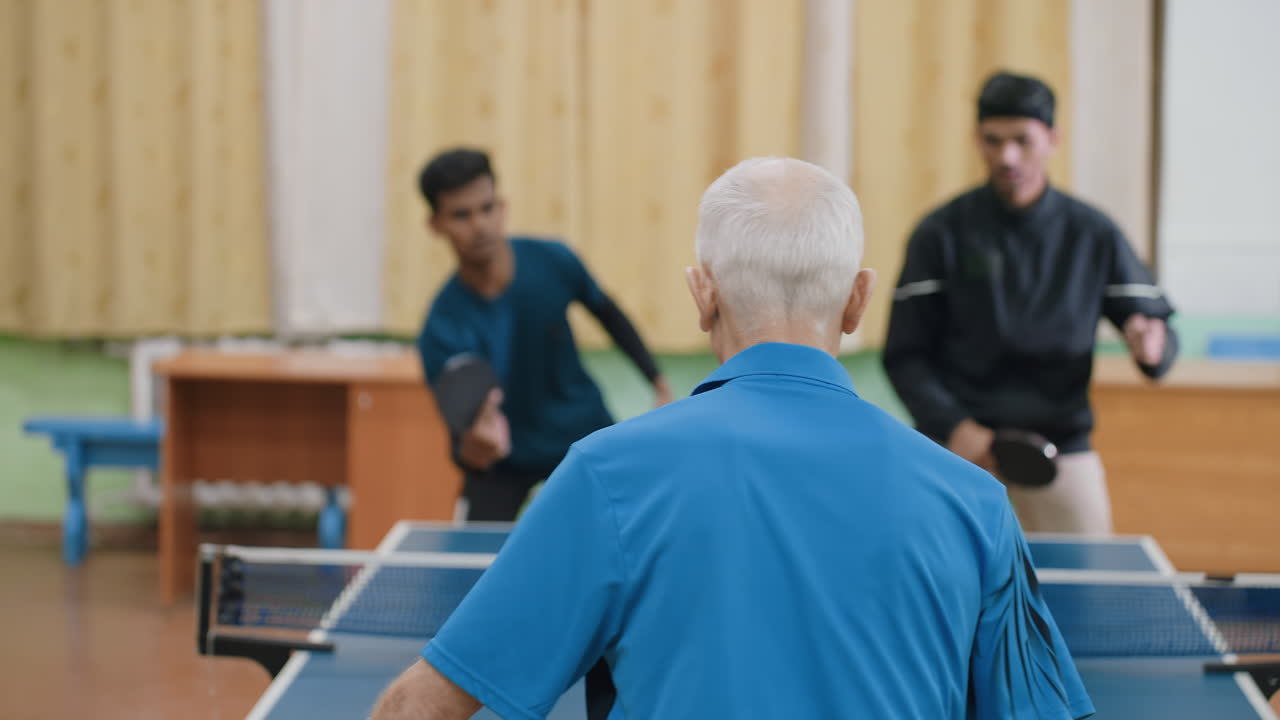 Senior athlete in blue shirt competes in indoor table tennis match against two young opponents, showing concentration, energy, and experience while background players appear slightly blurred