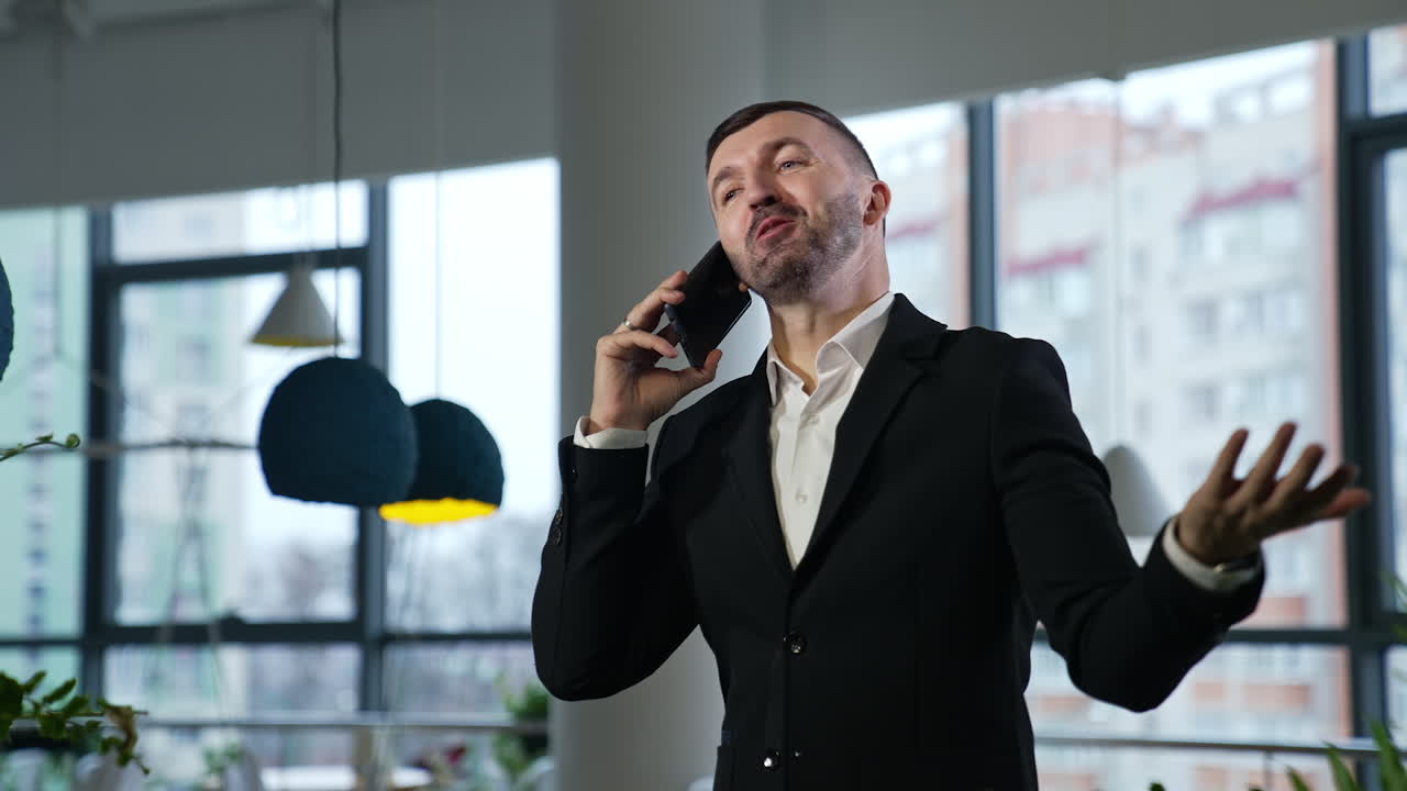 Entrepreneur wearing official suit in the office. Boss speaking on the smartphone and smiling. Windows in blur at the background.