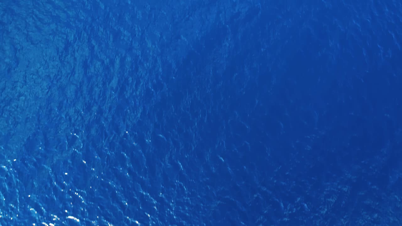 Drone daalt langzaam af naar diepblauw oceaanwater, gezien vanaf het oppervlak terwijl rimpelingen en golven over de top waaien, oceaantextuur.
