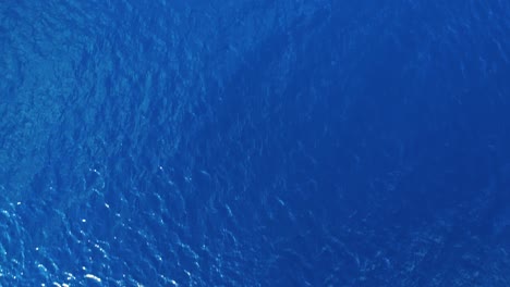 drone slowly lowers to deep blue ocean water view from surface as ripples and waves blow across top, ocean texture