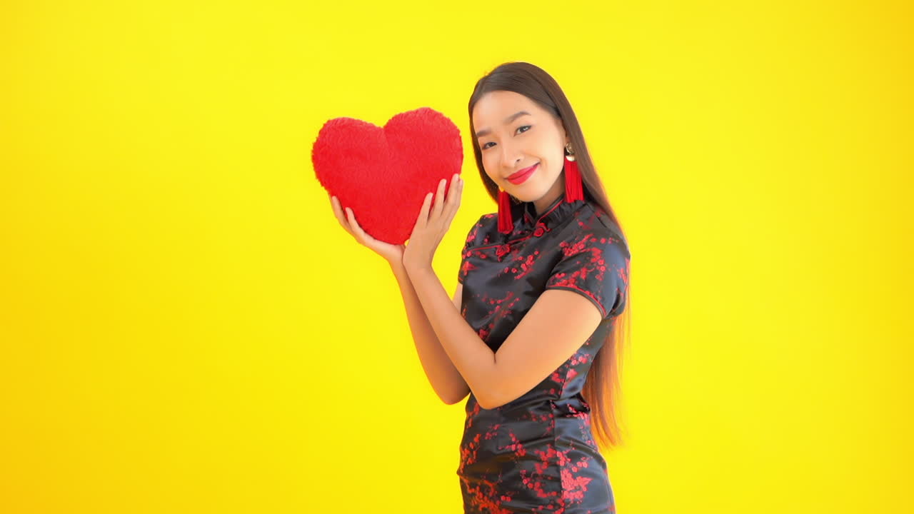 Chinese woman holding large red craft heart for Valentine's Day. Asian model posing with heart shaped pillow against yellow background. Smiling happy woman celebrating love. Copy space