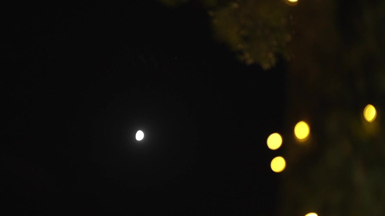 Luna luminosa nel cielo scuro con luci calde e sfocate che creano un effetto bokeh.