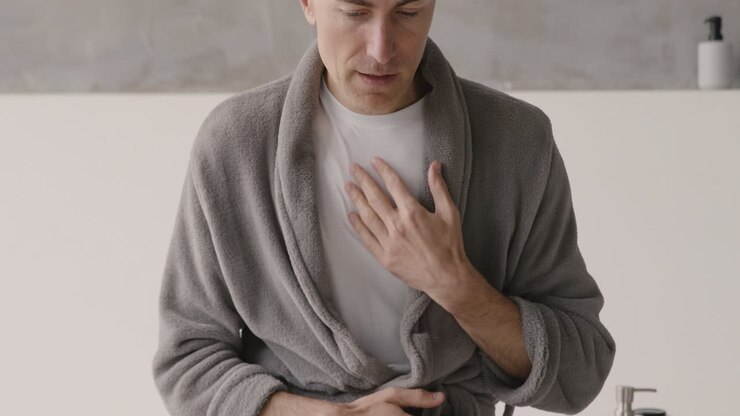 Uomo sofferente in abiti e vestaglia con dolore al petto che si tocca la zona del cuore al mattino a casa 1