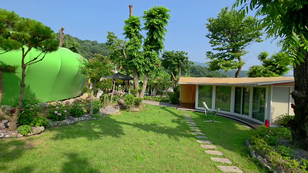 A contemporary glamping cabin and BBQ pavilion are nestled in a vibrant green garden with manicured trees and landscaped paths at Glamtree Resort in Gapyeong, South Korea