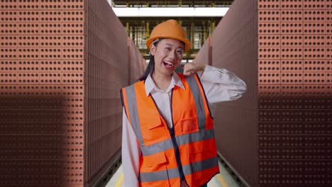 asian female engineer with safety helmet smiling to camera and making call me gesture while standing with red brick packed in stacks are stored