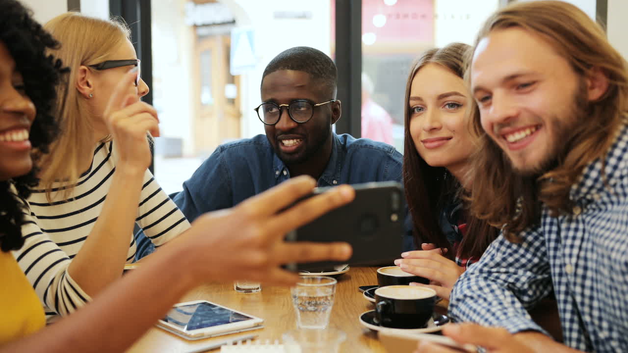 Close-up view of multiethnic group of friends talking and watching a video on a smartphone sitting at a table in a cafe