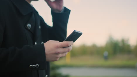 close up of individual in black coat multitasking by operating phone and sipping tea outdoors, showcasing focus and modern lifestyle in urban environment with blurred background and moving car