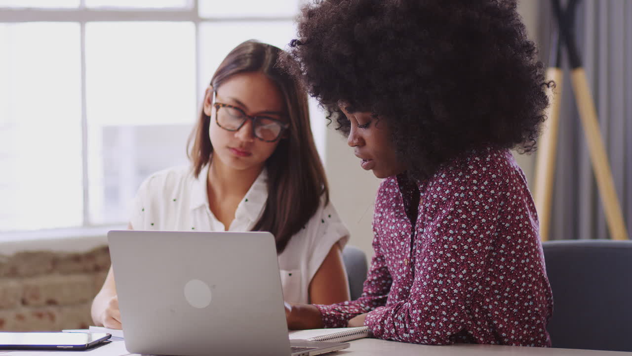 Two millennial female business creatives talking and making notes in a team meeting, close up
