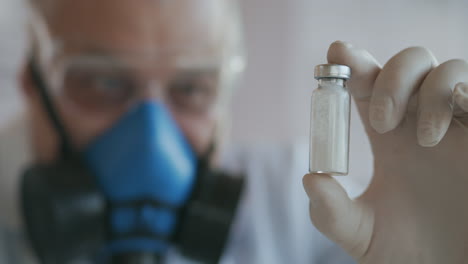 Primer-Plano-Extremo-De-Un-Científico-Con-Un-Respirador-Azul-Y-Gafas-Protectoras-Que-Desarrolló-Una-Vacuna-Contra-El-Coronavirus-Sosteniendo-Una-Ampolla-De-Polvo-Blanco.-Sustancias-Narcóticas-Y-Analgésicos.-Imágenes-4k-De-Alta-Calidad