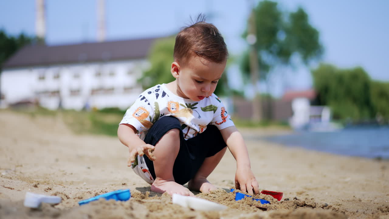 Cute little Caucasian boy plays in the sand with toys. Focused kid takes wet sand and digs it with his shovel. Child plays at shore.
