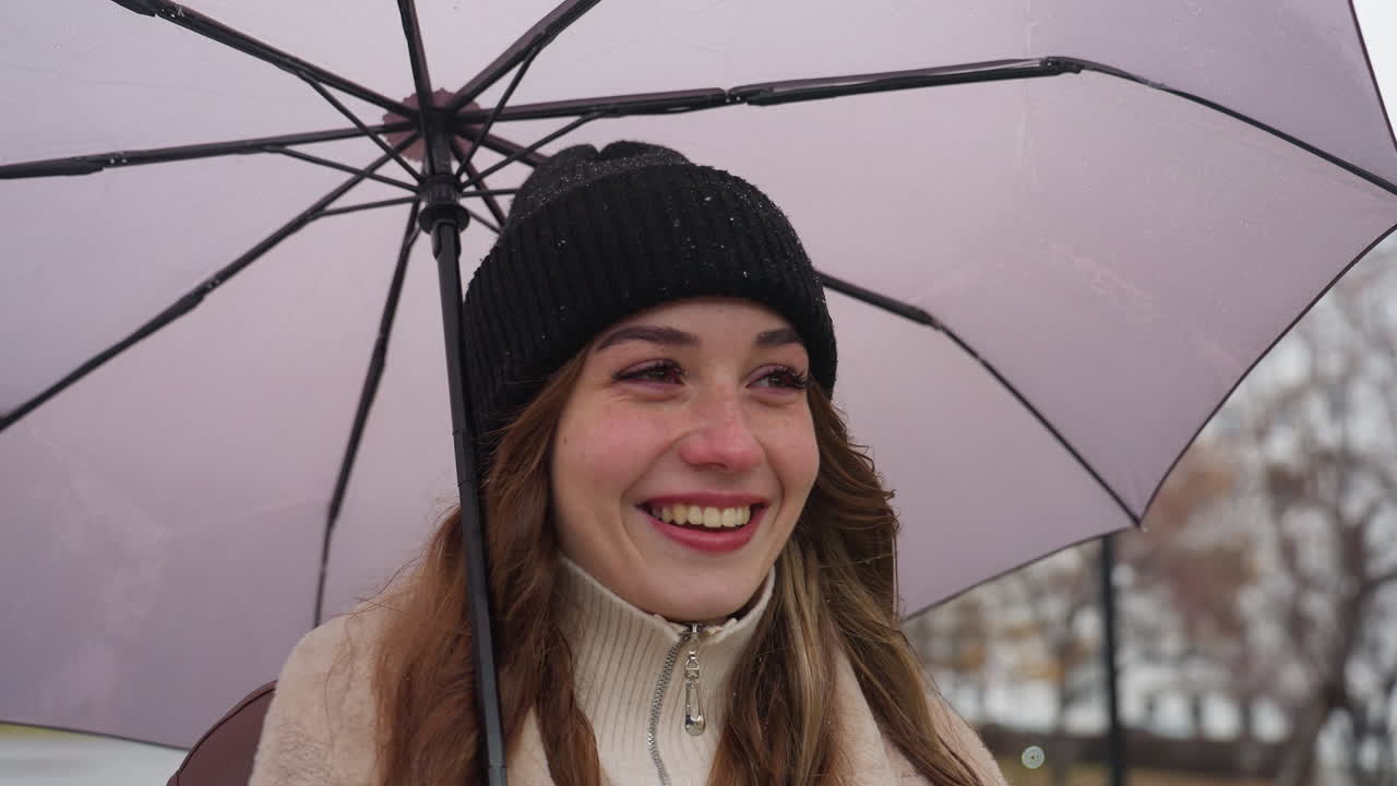 Smiling student holding umbrella, wearing black knit cap and brown shearling jacket, enjoying cold overcast day with light snowfall, bright happy expression, winter weather, peaceful atmosphere