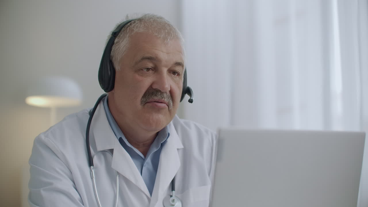 un médico anciano vestido con túnica blanca y con un estetoscopio en el cuello está consultando a un paciente en línea utilizando la tecnología de videollamada en una computadora portátil
