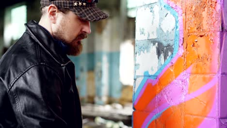 bearded guy in casual clothing is drawing abstract image with aerosol paint on pillar inside old building. abandoned house, youth subculture and creativity concept.