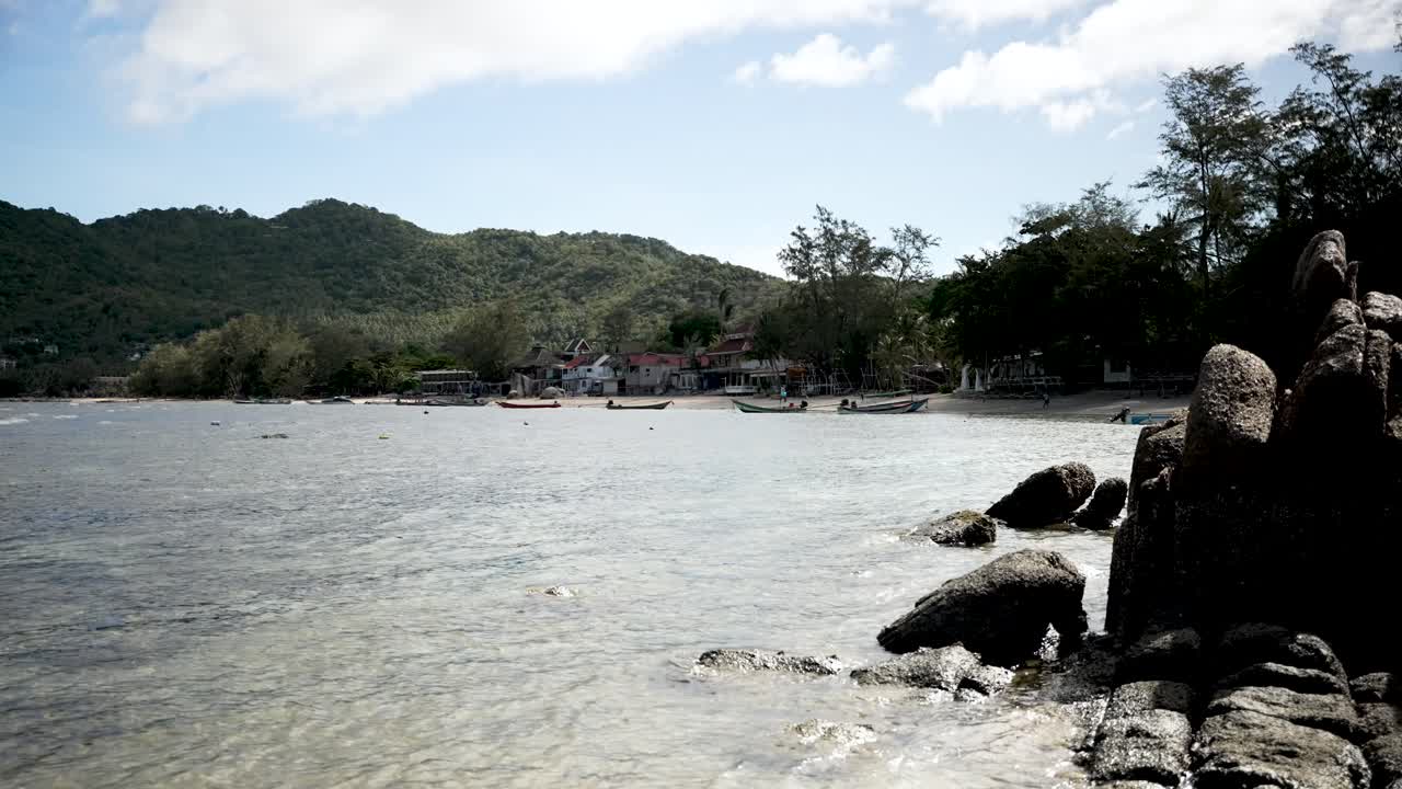 부드러운 파도, 물 위에 떠다니는 작은 배, 그리고 배경에 울창한 초록색 산이있는 조용한 열대 해변, 편안한 탈출을 위해 완벽합니다.