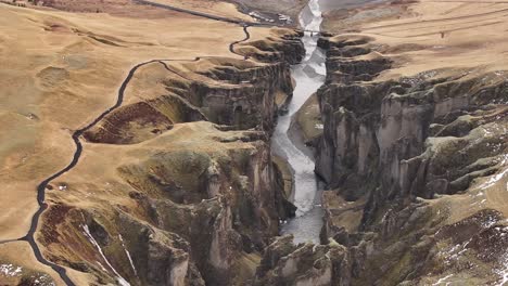 aerial view of fjaðrárgljúfur canyon in iceland, with steep cliffs and a meandering river cutting through the rugged golden landscape under soft daylight.