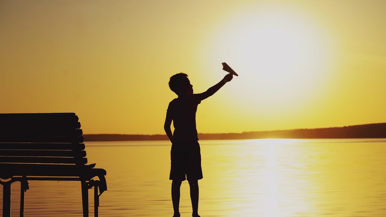 Happy child is playing with a paper airplane. Boy on a wooden bridge on the river in a picturesque setting at sunset.
