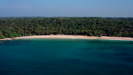 Lateral drone flyby from left to right along a tropical beach with turquoise waters and palm forest at Silent Beach, Sri Lanka during midday.